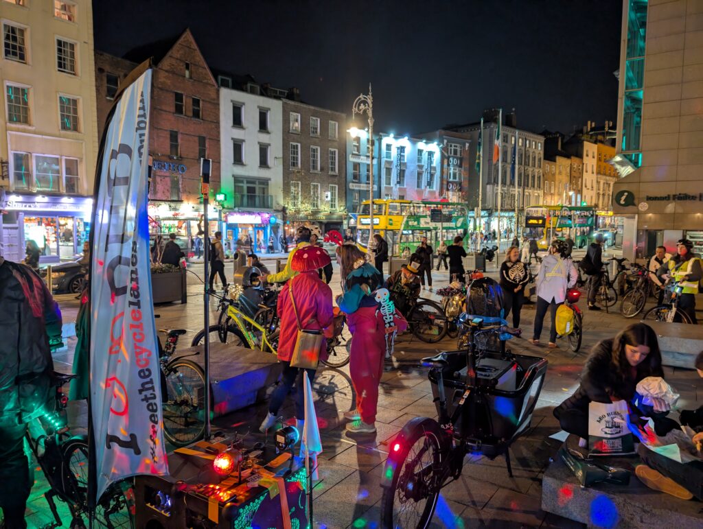 People gathered with their bicycles in a pedestrian city square. Some are dressed up in costumes, there are coloured lights and flags.