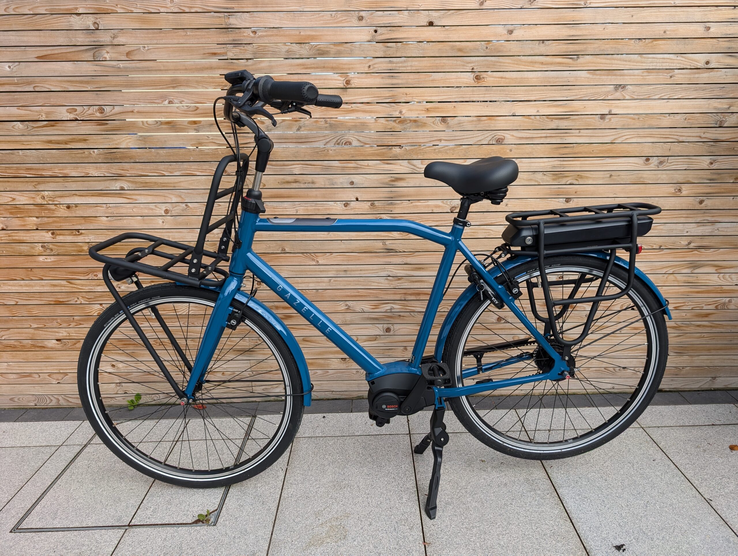 Blue bicycle in front of a wooden fence. The bicycle has a front and rear racks for carrying cargo, and is an electric bike.